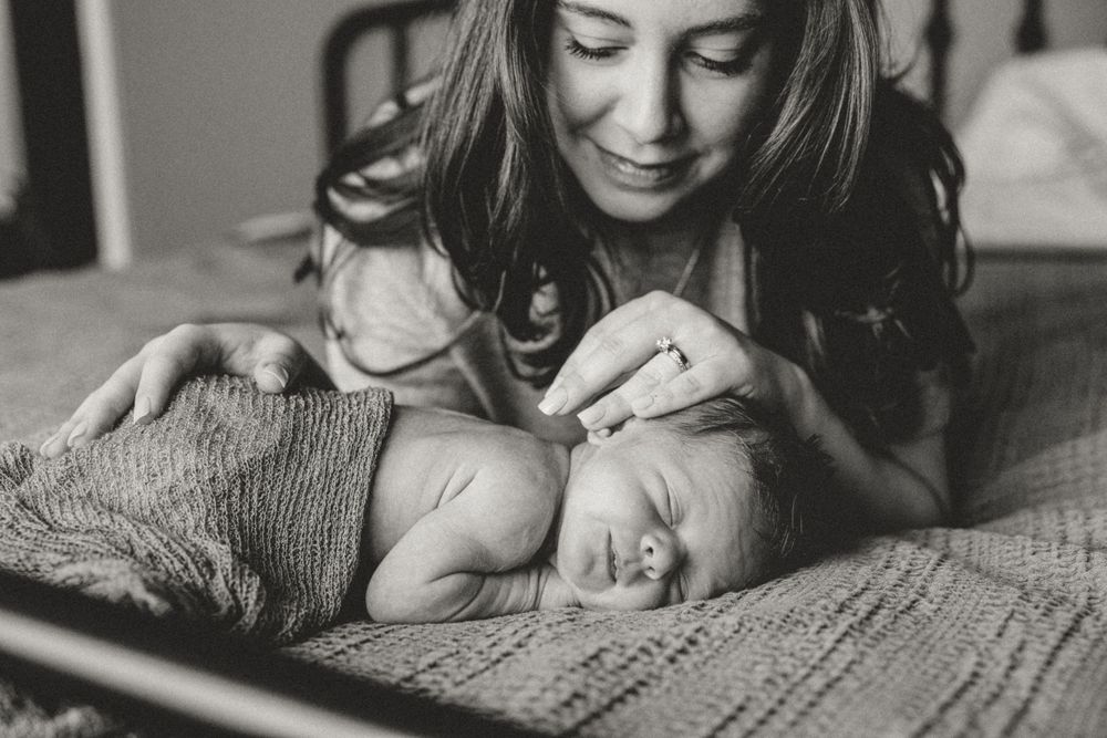 Black and white portrait of mother tenderly watching sleeping newborn baby wrapped in knit blanket on bed.