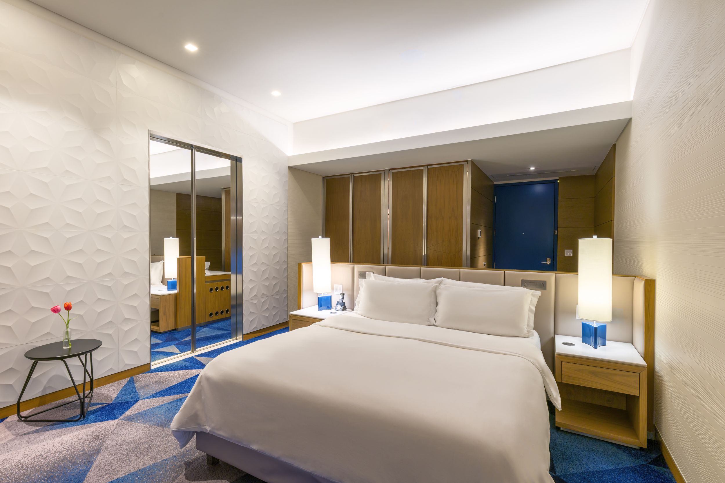Modern hotel room interior with white bed, wooden accents, blue carpet, and contemporary lighting fixtures.