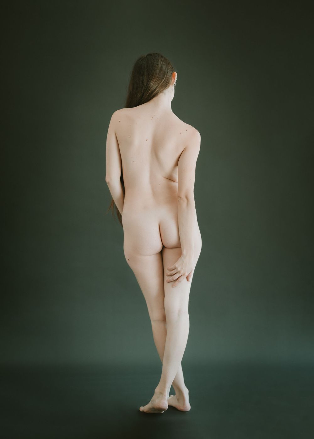 Artistic nude studio photograph showing a figure from behind against a dark green background.