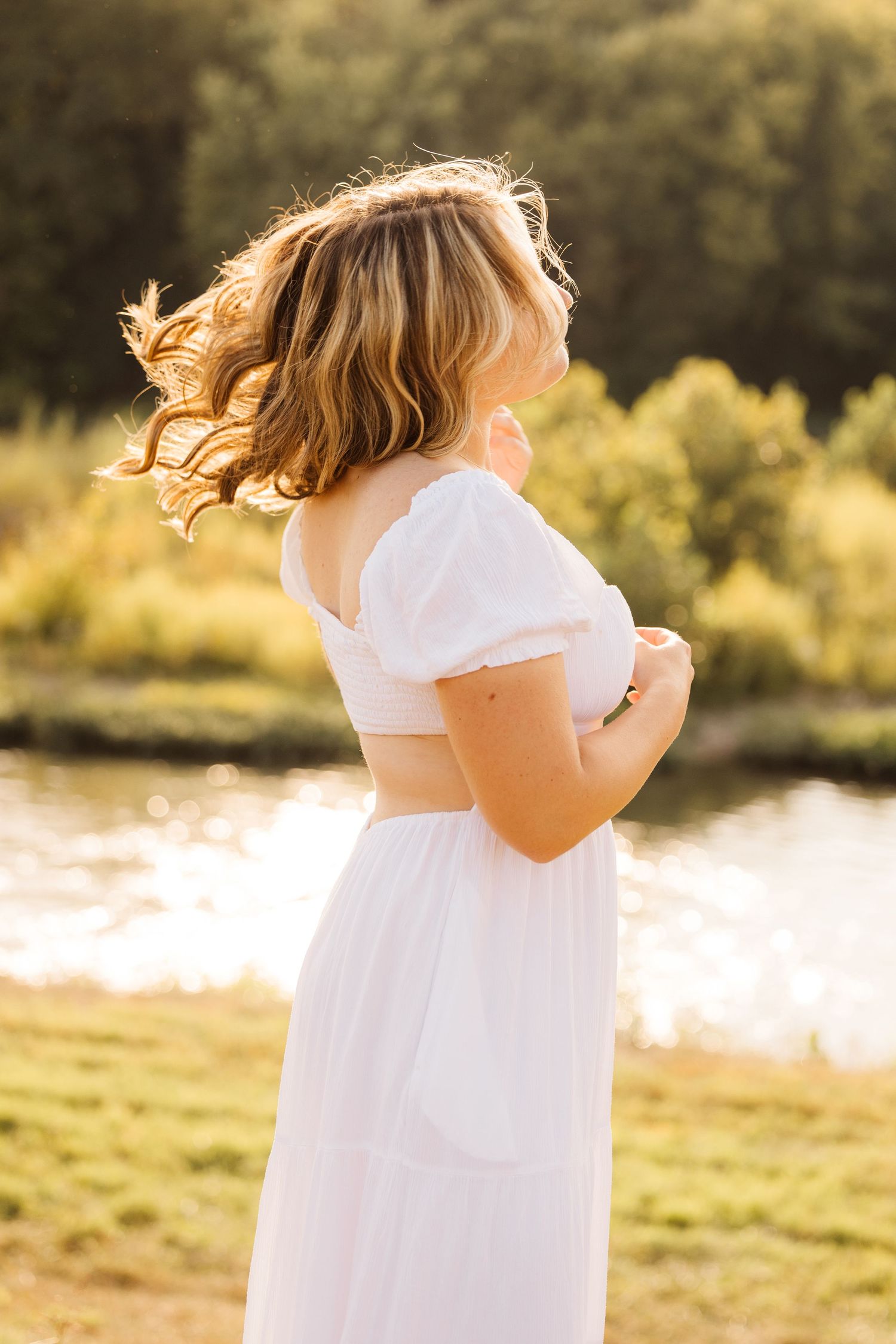 Person in a flowing white outfit twirls at golden hour by a sunlit lake, with their blonde hair catching the light.