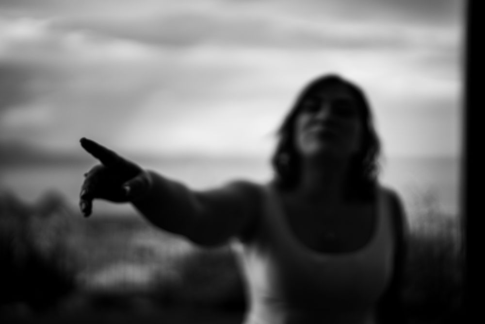 Blurred black and white silhouette of person in tank top pointing into distance against cloudy sky.