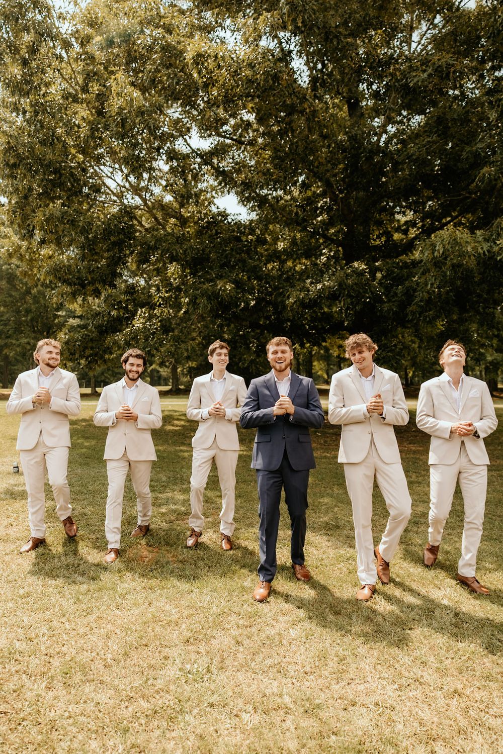 A group of groomsmen in light beige suits stand together outdoors with one in navy blue at a wedding celebration.