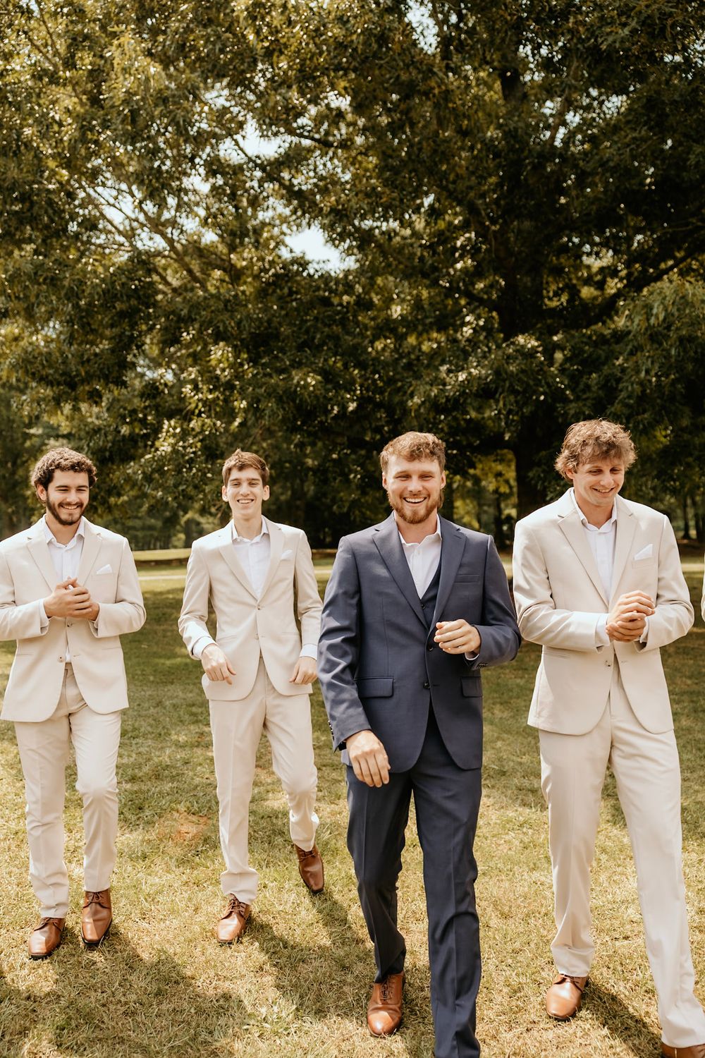 Four groomsmen stand together outdoors wearing suits, with three in light beige and one in navy blue in a garden setting.