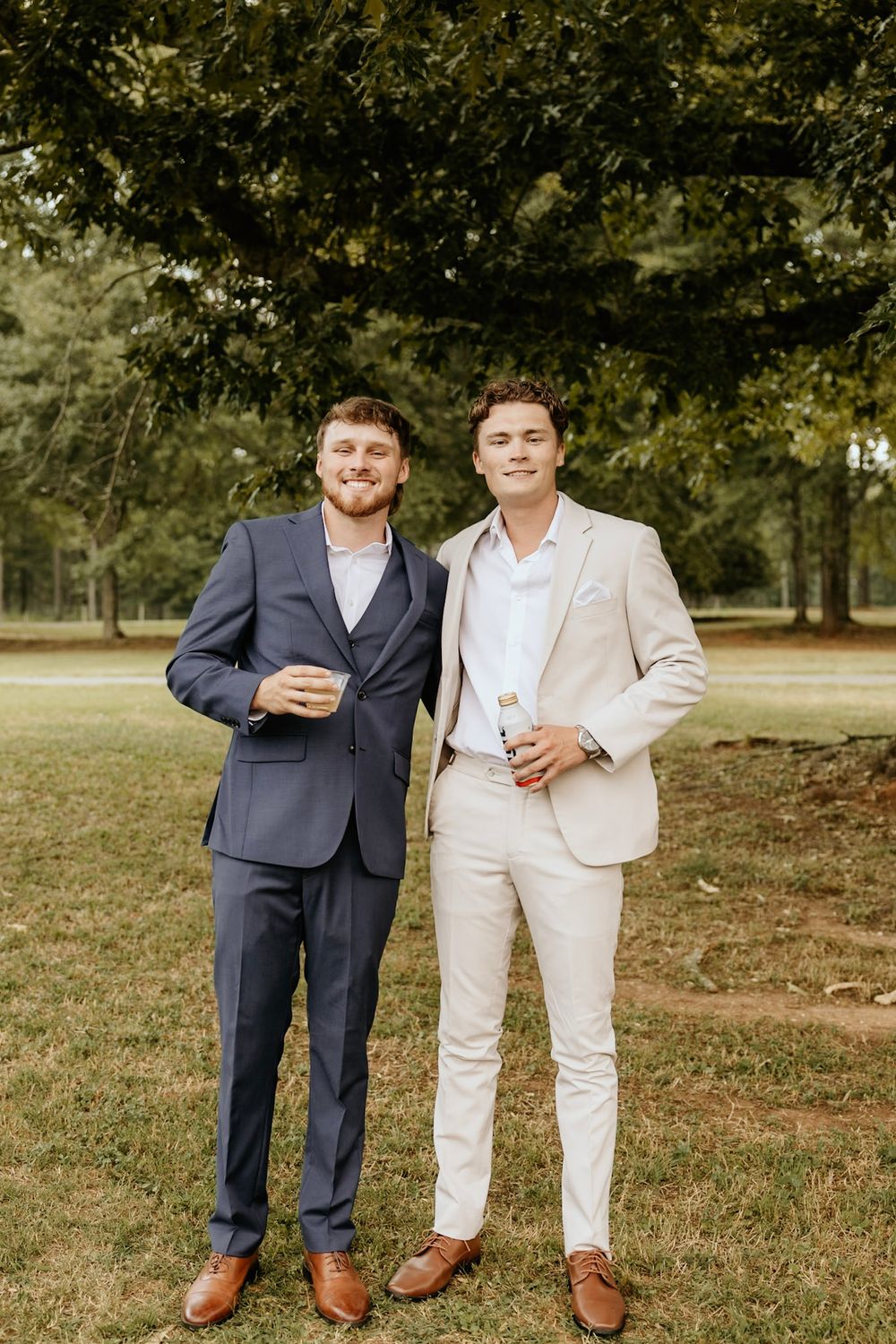 Two people in formal suits, one in navy and one in beige, standing together outdoors on grass with trees in background.