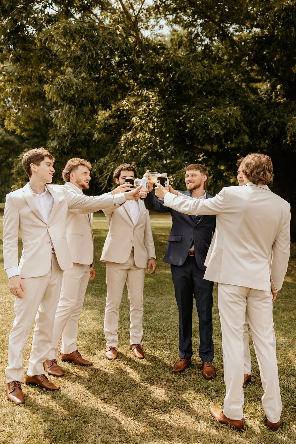 Groomsmen in light beige suits toast with drinks at an outdoor wedding celebration on a sunny day.