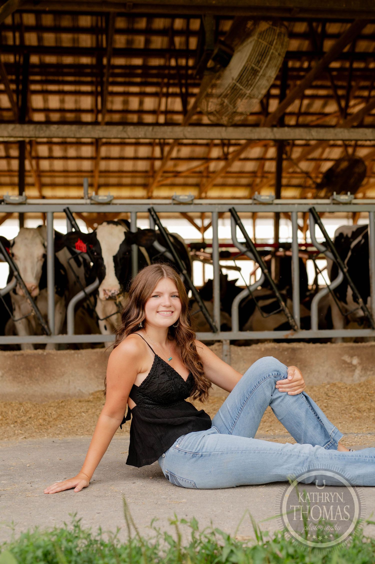 Casual portrait of someone relaxing on the ground in a black top and jeans inside a dairy barn.