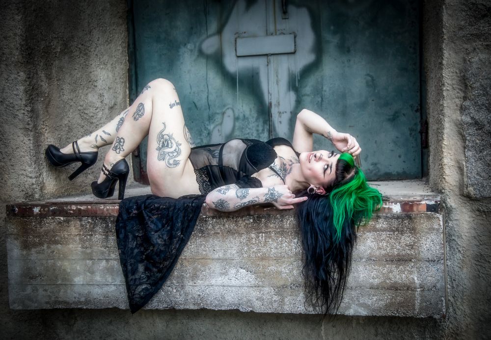 Tattooed woman with green hair posing on a ledge in black lingerie and heels against a grungy wall.
