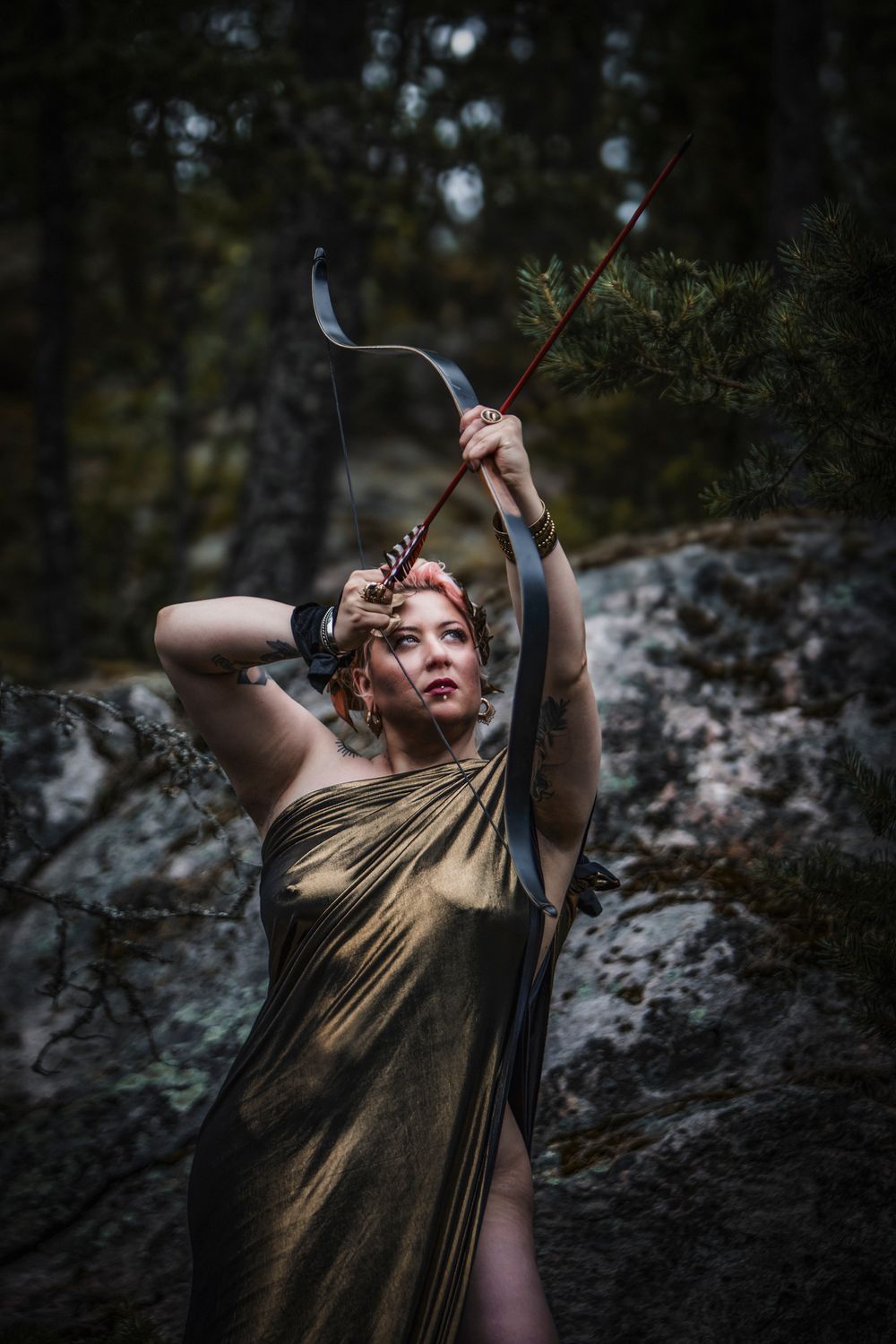 Woman dressed as Greek goddess Artemis in gold draped gown, drawing bow and arrow in mystical forest setting.