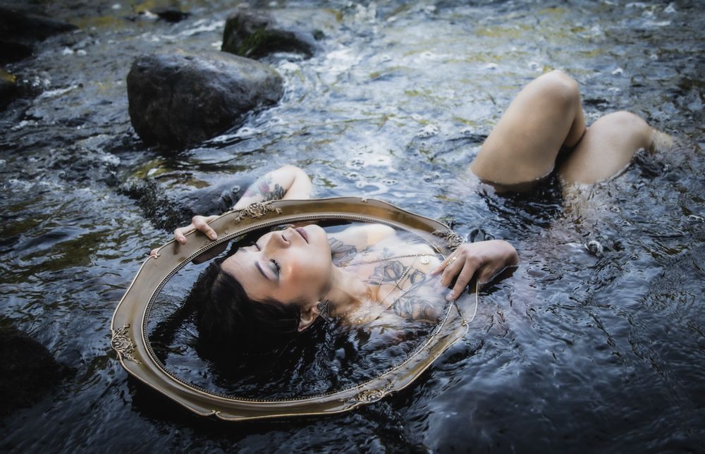 Tattooed woman lying in a rocky stream holding an ornate mirror, fine art photography.