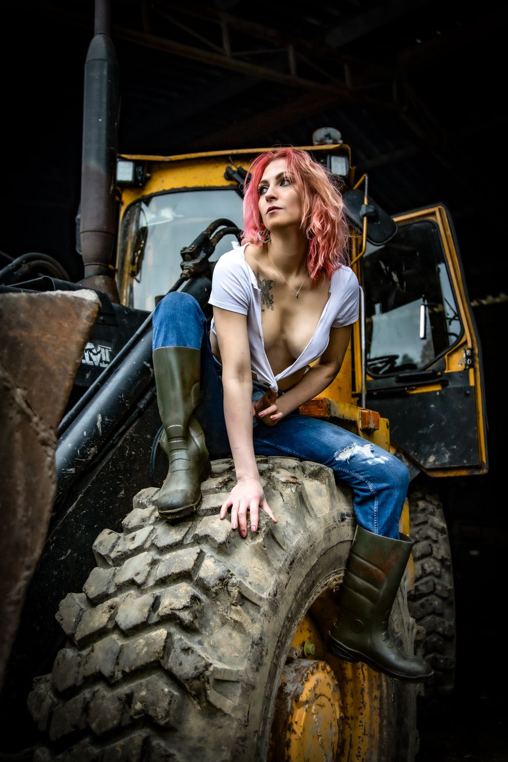 Model with pink hair poses on large industrial tire wearing boots and casual attire in gritty setting.