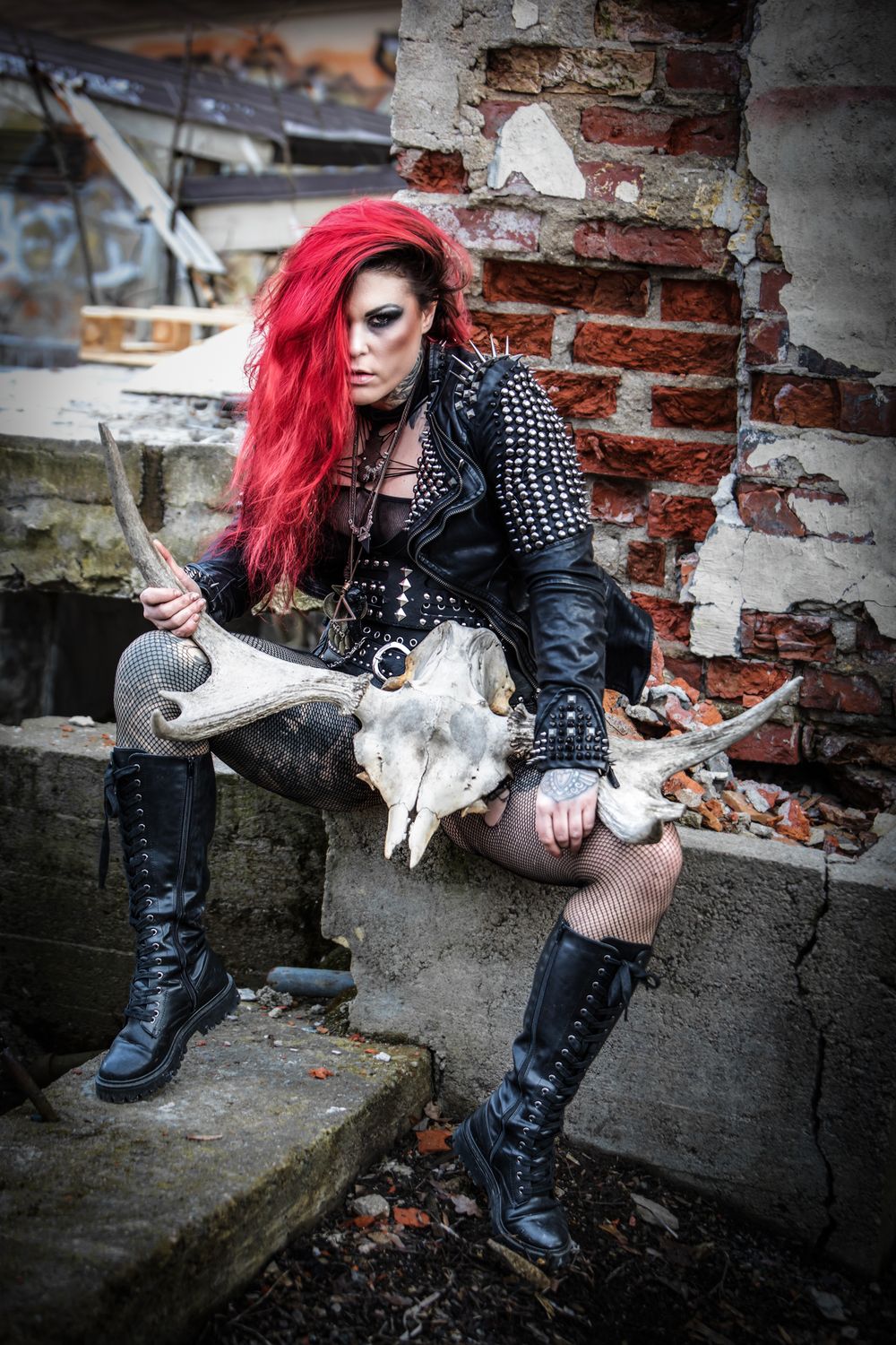 Gothic styled figure in black leather and spikes sits on concrete steps with animal skull in post-apocalyptic setting.