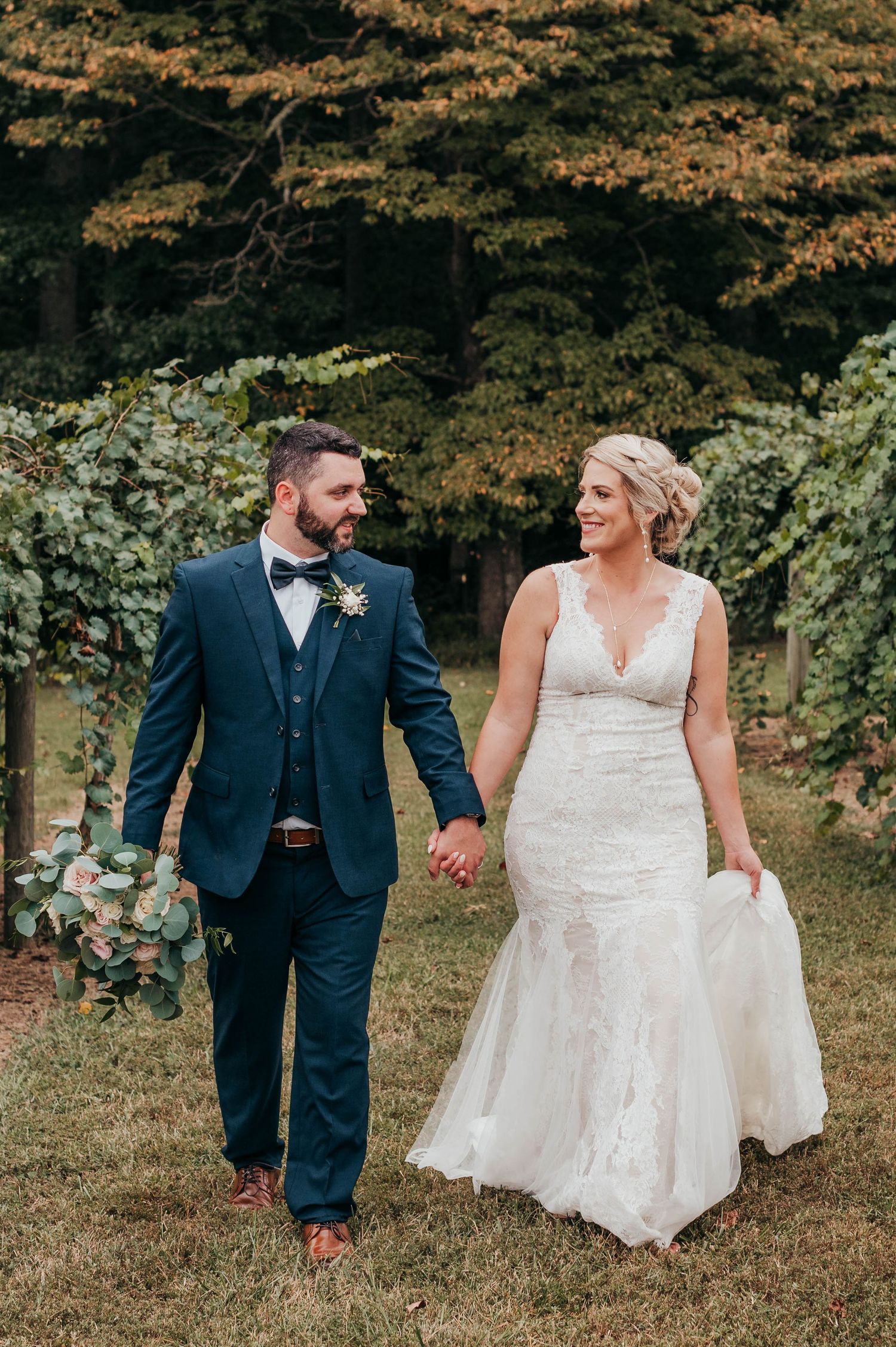 A couple walks hand in hand outdoors, with the bride in a white lace gown and groom wearing a navy suit with bowtie.