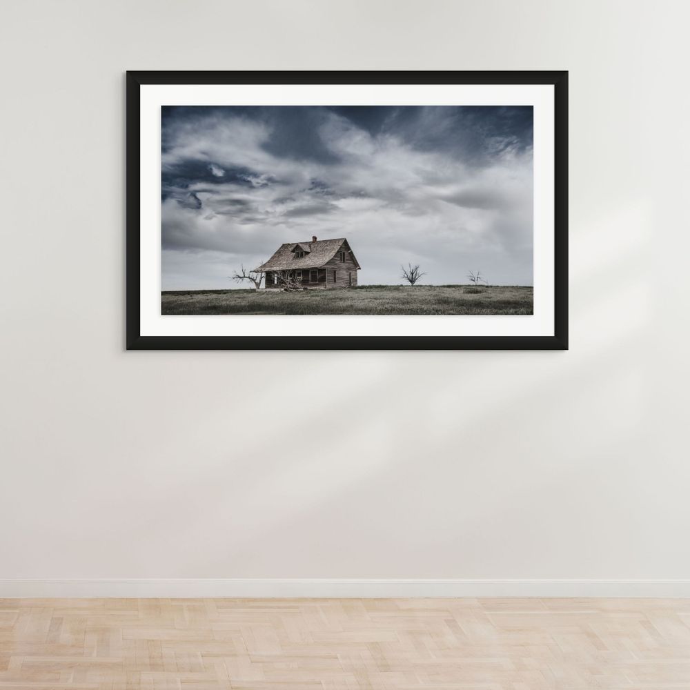 Framed photograph of an abandoned farmhouse standing solitary on the prairie under dramatic skies.