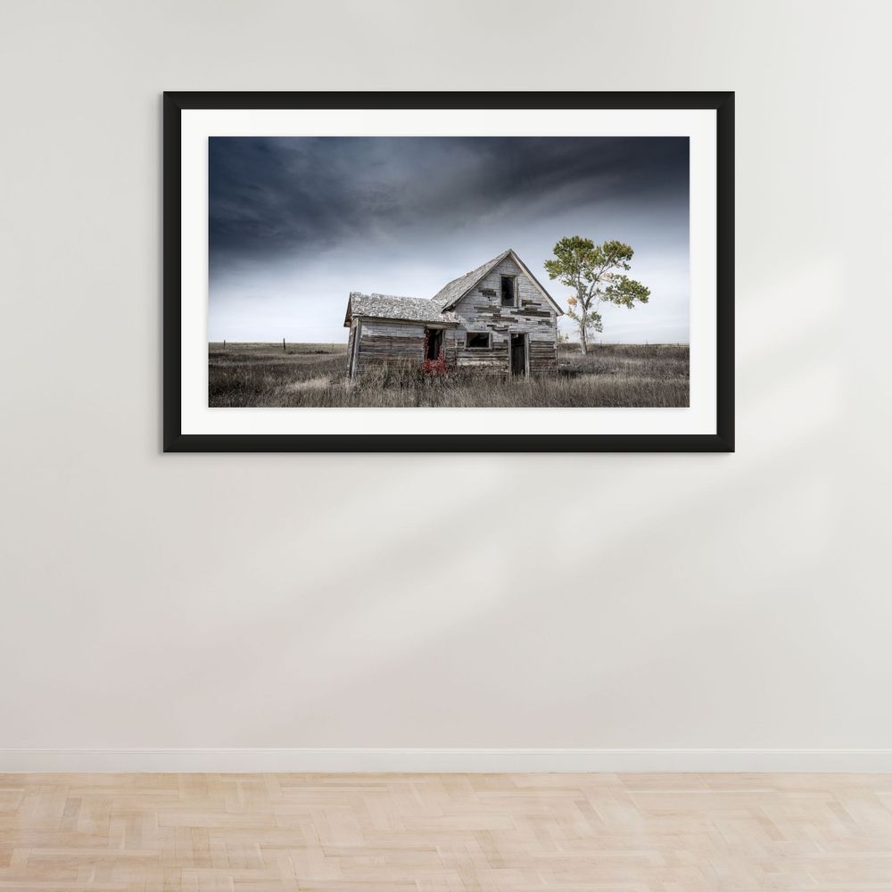 Weathered abandoned house with peeling paint stands alone on prairie landscape in black frame.