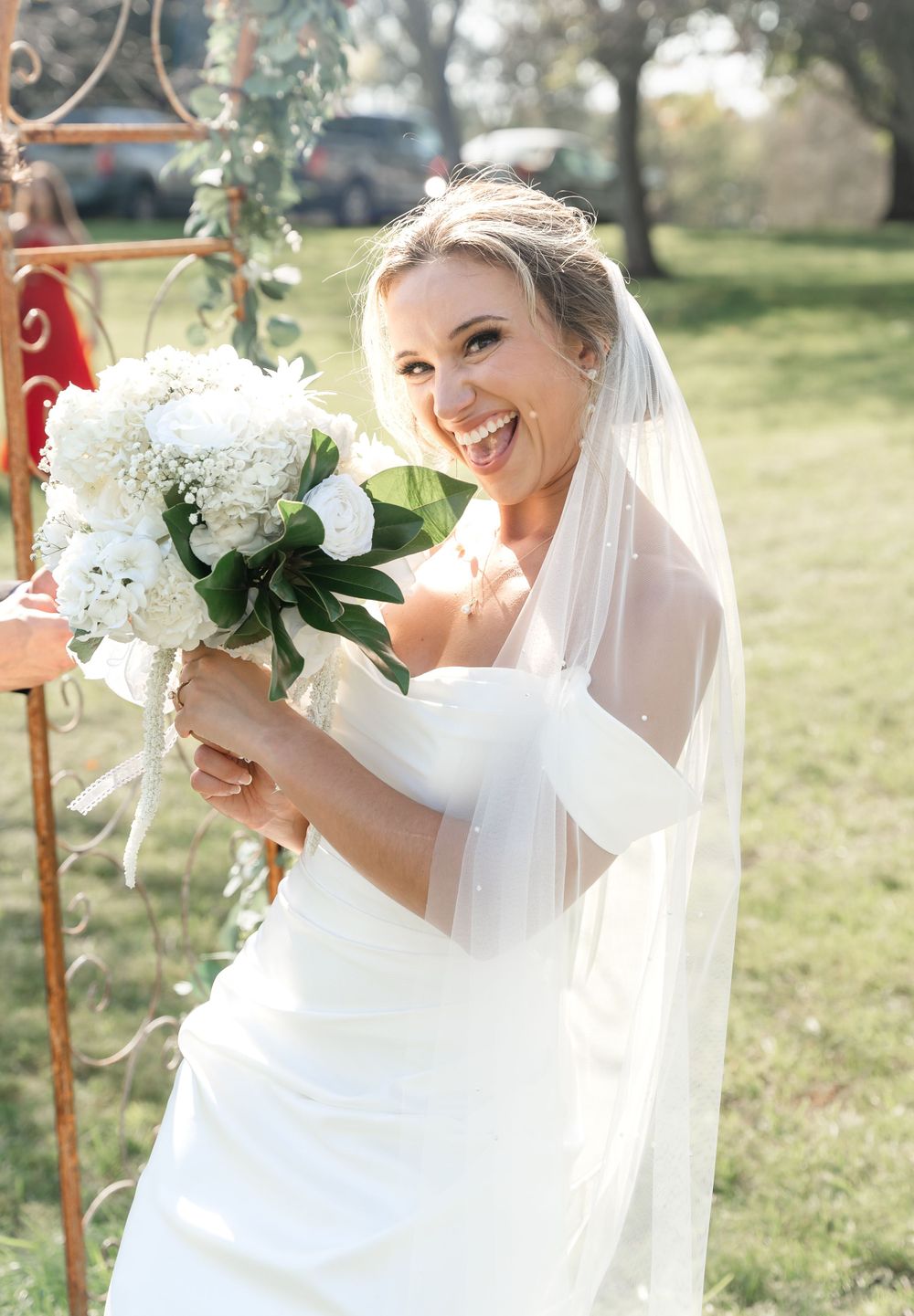 Joyful blonde bride in white gown and veil holding white hydrangea bouquet outdoors on a sunny wedding day.