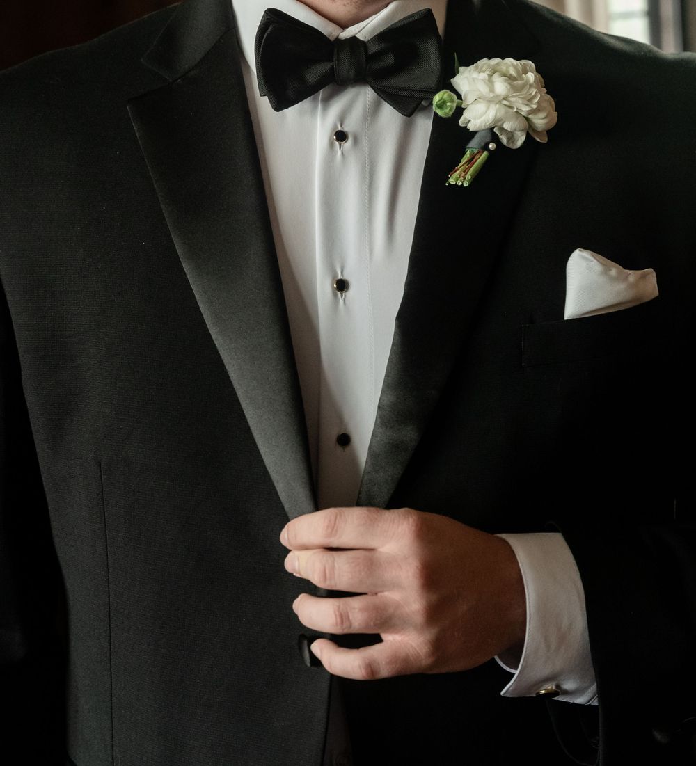 Groom in classic black tuxedo with bow tie and white rose boutonniere adjusting his jacket lapel.