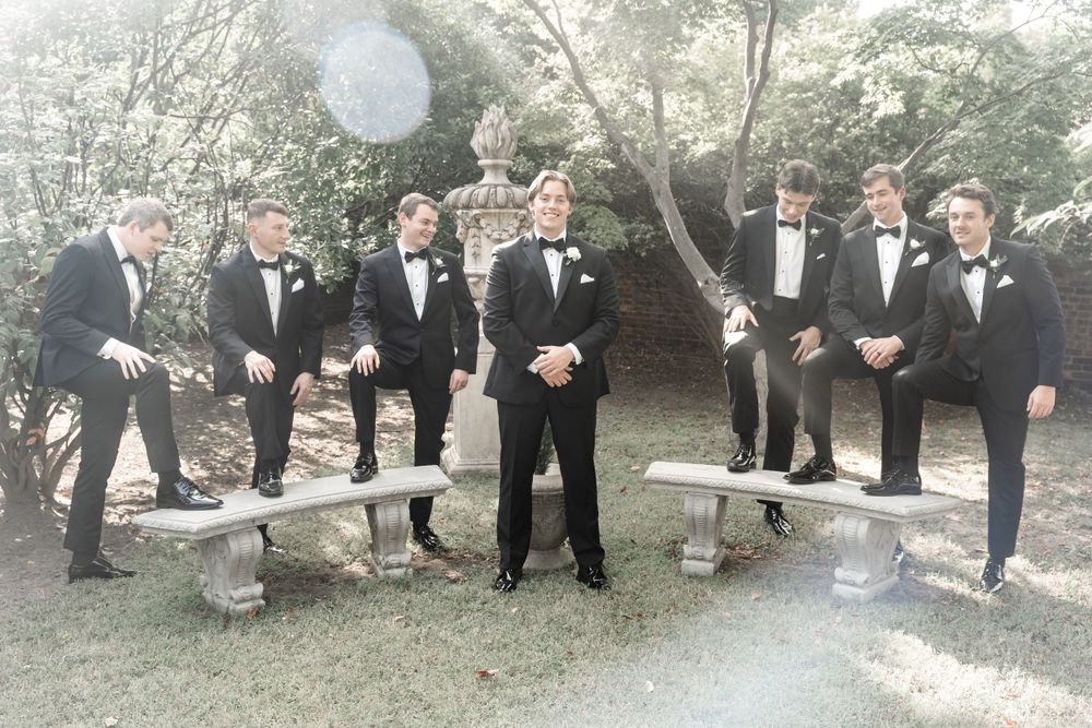 Groom and groomsmen in matching black tuxedos posing casually in a lush garden setting with stone benches.