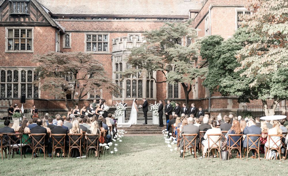 Outdoor wedding ceremony on lush lawn of historic brick manor with seated guests.
