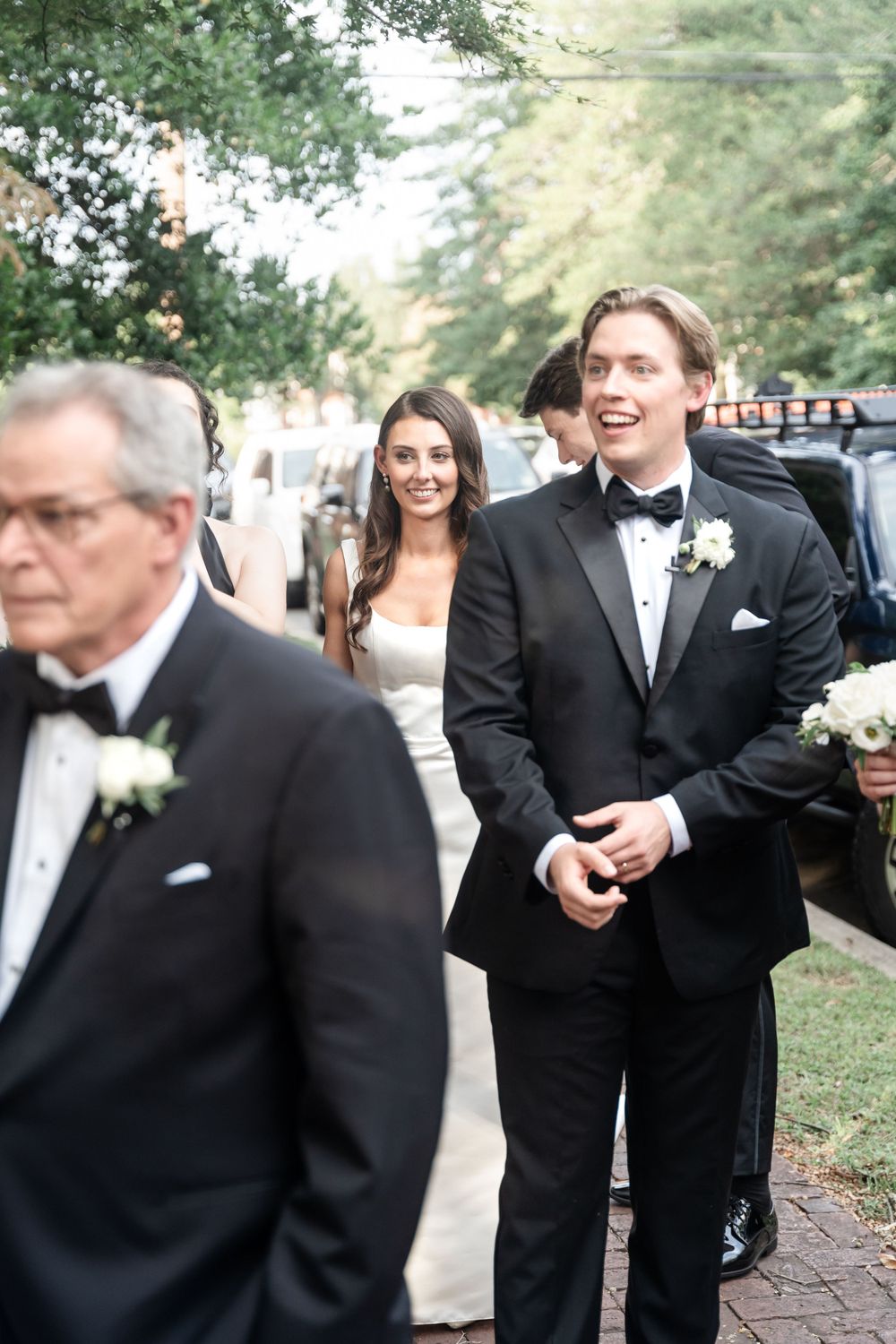 Bride in white gown walking with groomsmen in black tuxedos along a tree-lined street before the ceremony.