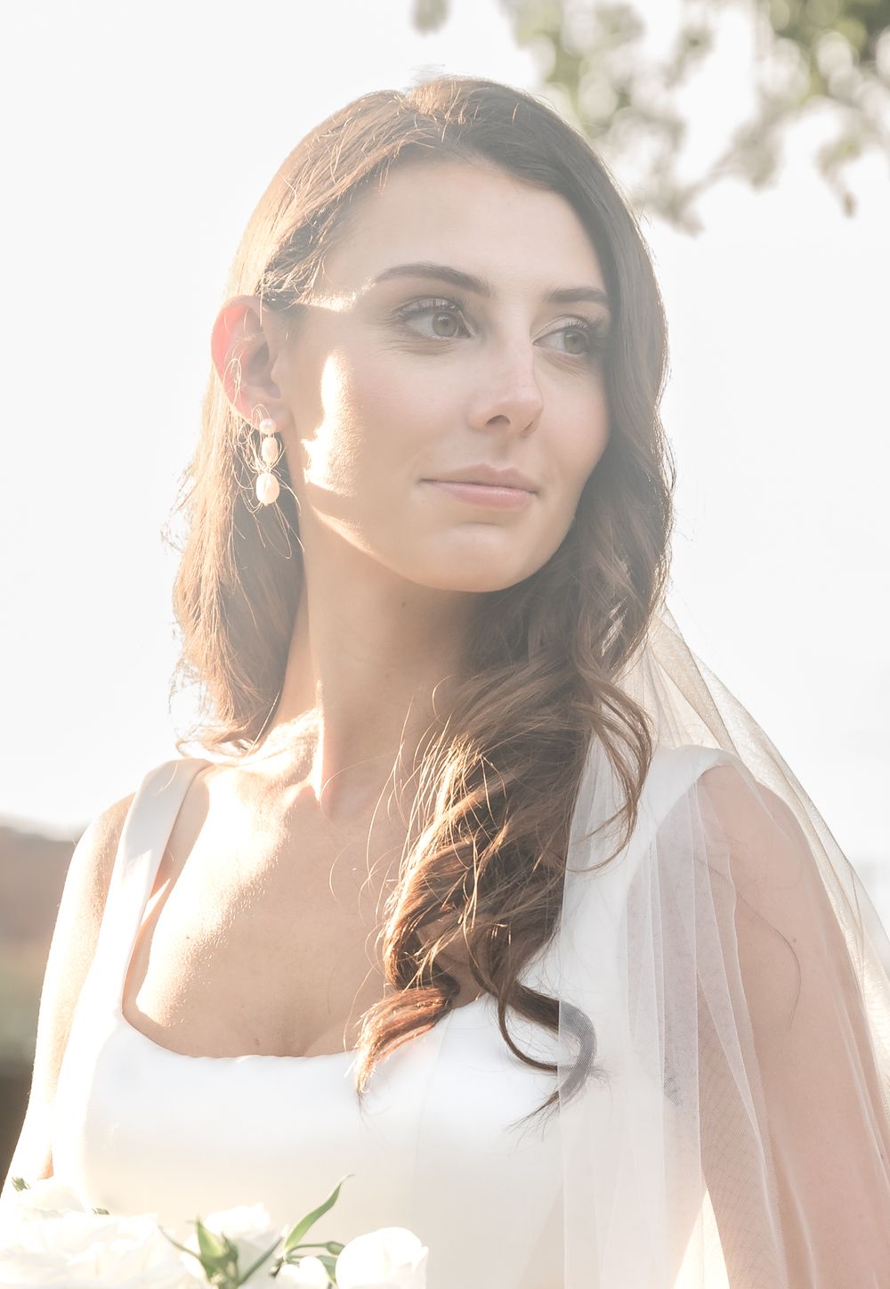 Elegant bride in white gown with veil and drop earrings, soft golden backlight.