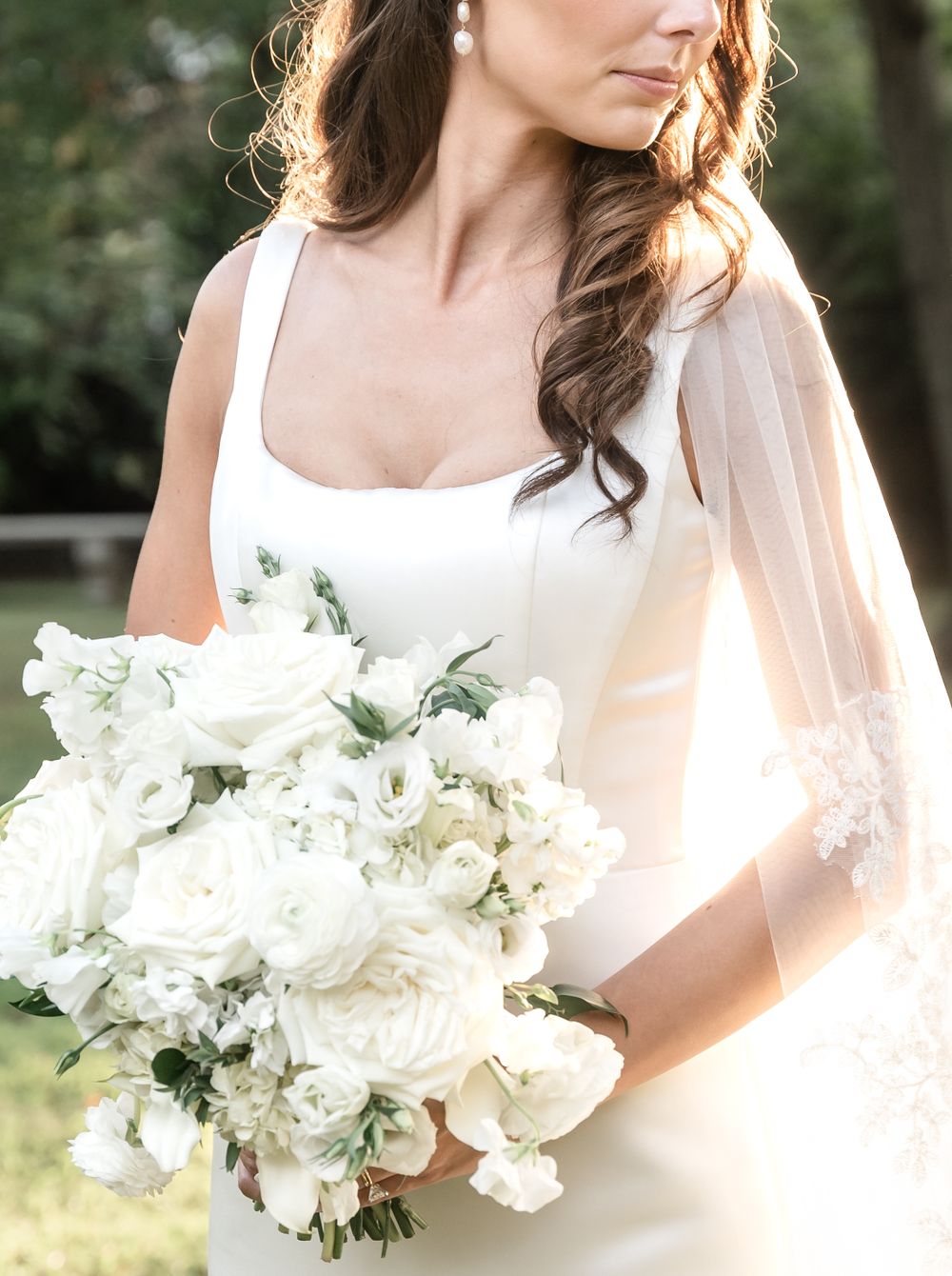 Bride in sleek white gown holding lush all-white bouquet of roses and ranunculus.