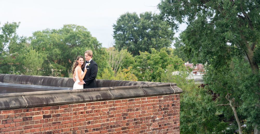 Bride and groom embracing on rooftop brick parapet surrounded by green trees.
