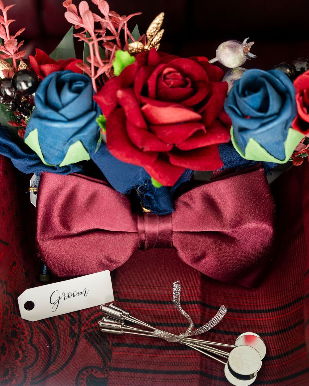Groom wedding flat lay with red and blue roses, burgundy bow tie, and cufflinks.