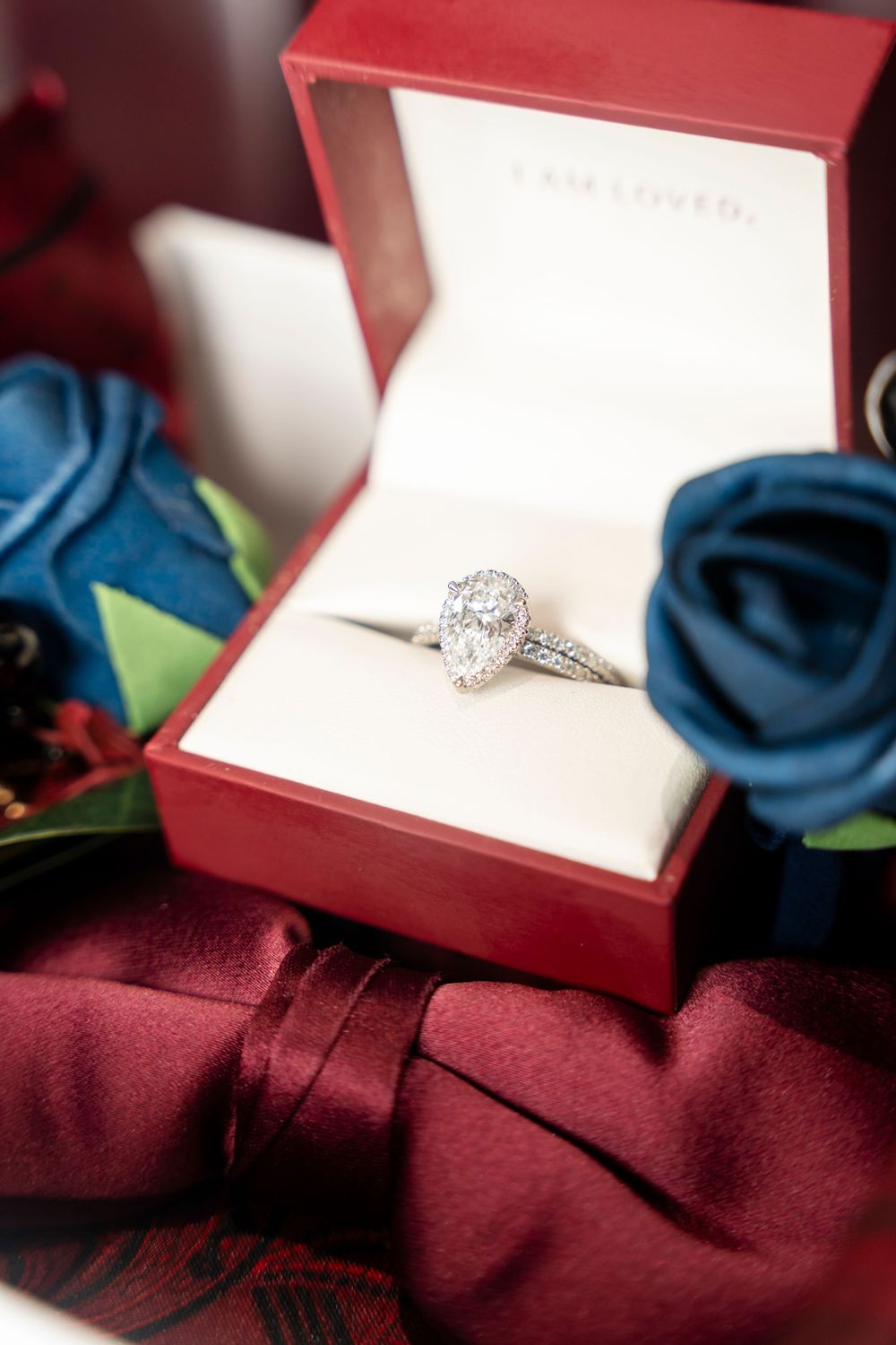 Diamond halo engagement ring in red box surrounded by blue and red fabric roses.