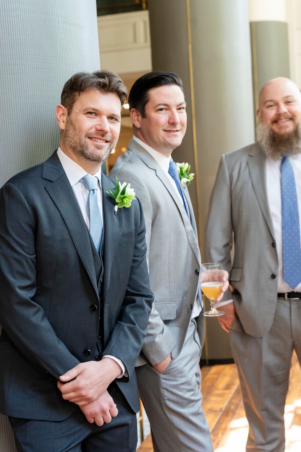 Three groomsmen in suits with boutonnieres smiling at a wedding reception venue.