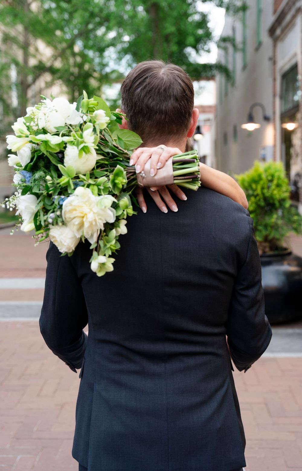 Groom in black suit embraced by bride holding large white and green bouquet, photographed from behind outdoors.