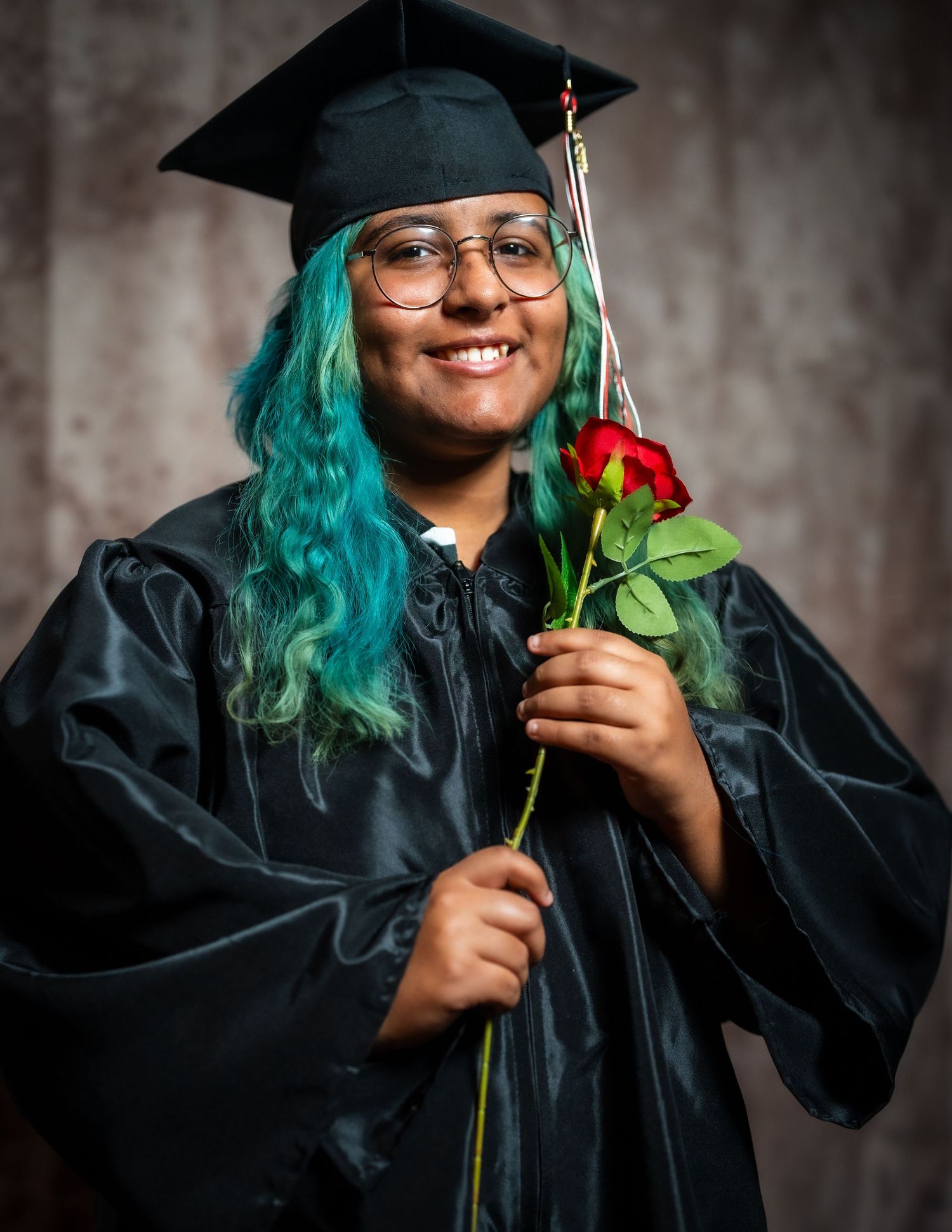 Graduate in black cap and gown with teal hair holds red rose while smiling.