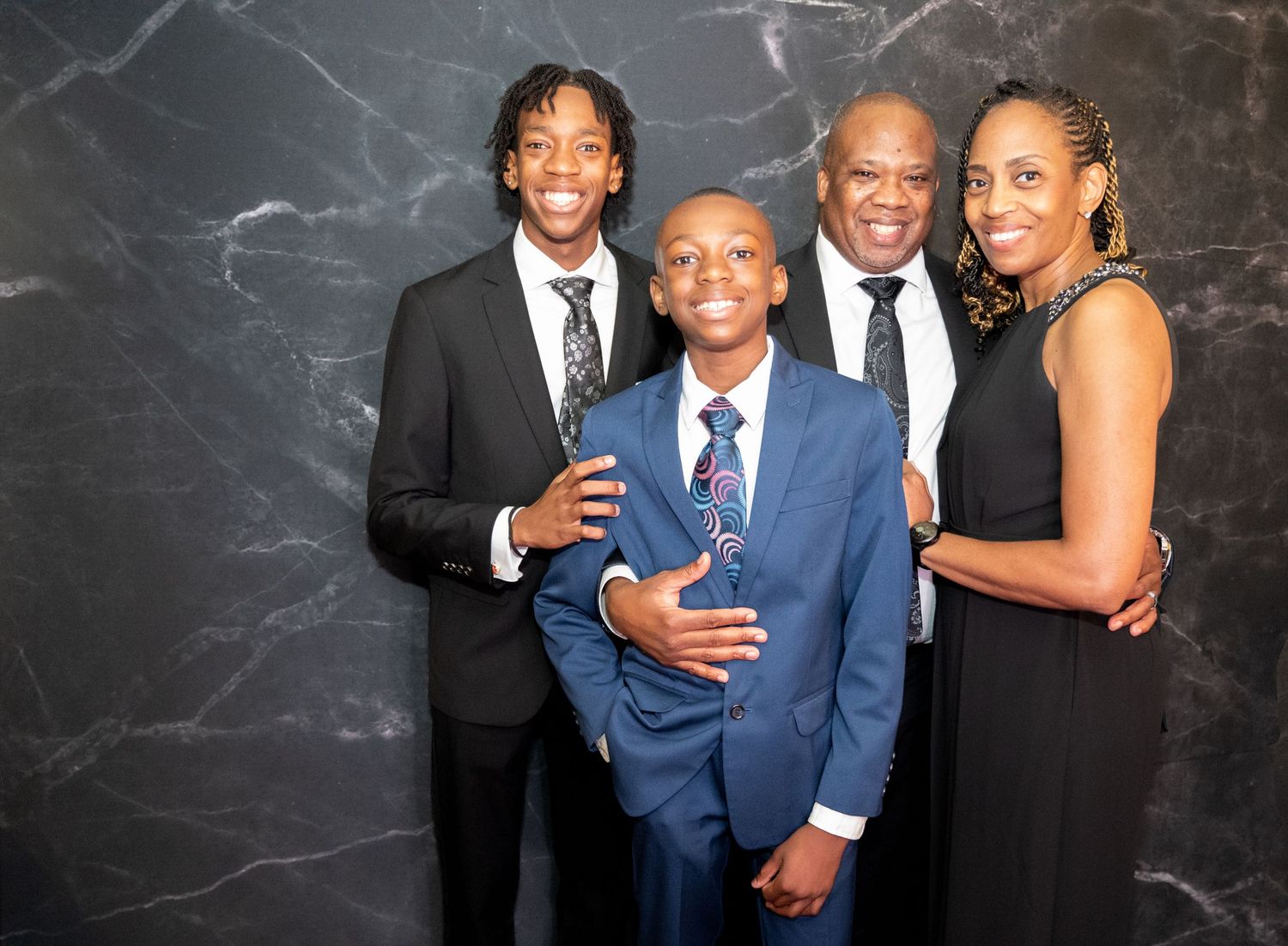 A formal family photo of four people dressed in elegant attire against a dark marbled background.