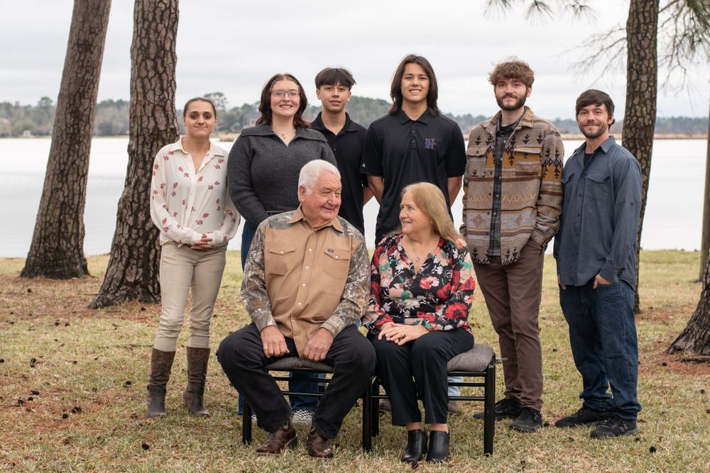 Group photo outdoors with several people standing behind two seated individuals, beside pine trees and a lake in background.