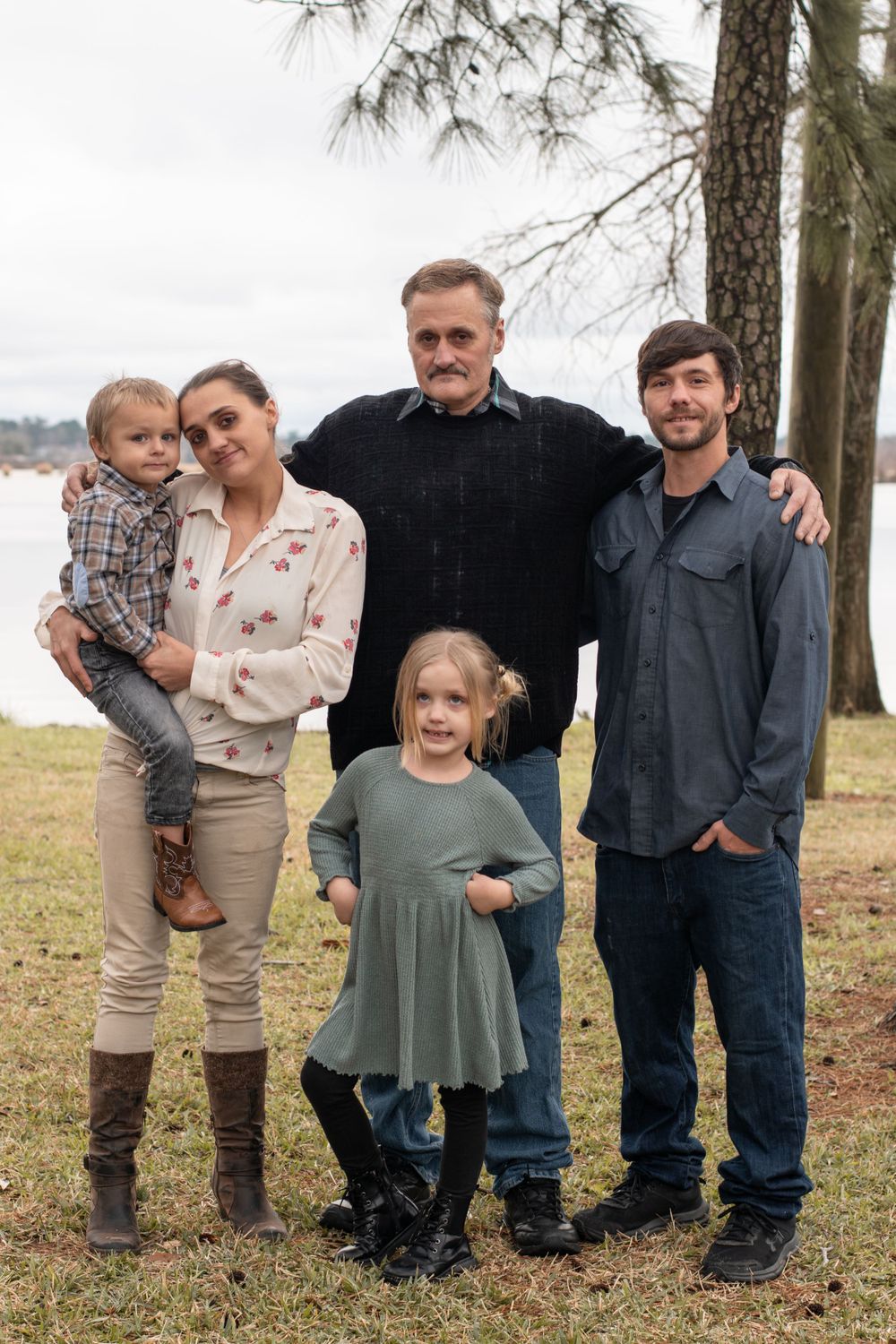Family group of five posing together outdoors near pine trees on a cloudy day by the water.