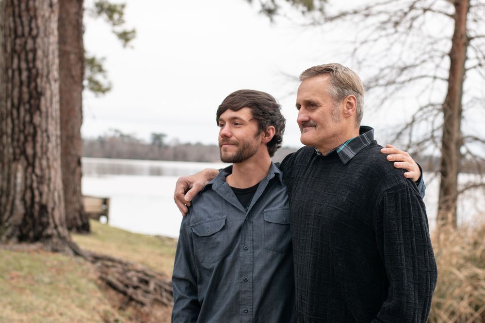Two people wearing dark jackets stand together beside a lake with tall pine trees in the background.