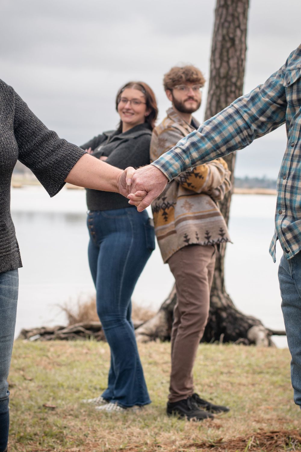 A group holds hands in a circle by a lake, with a tree trunk visible in the background on an overcast day.
