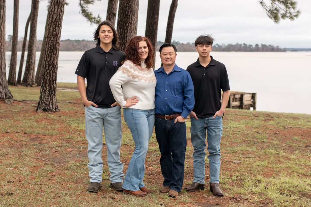 People pose together outdoors by a lake with tall pine trees in the background.
