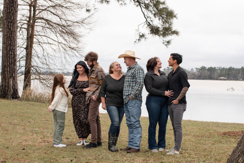 A group of friends laughing together by a scenic lake surrounded by pine trees on an overcast day.
