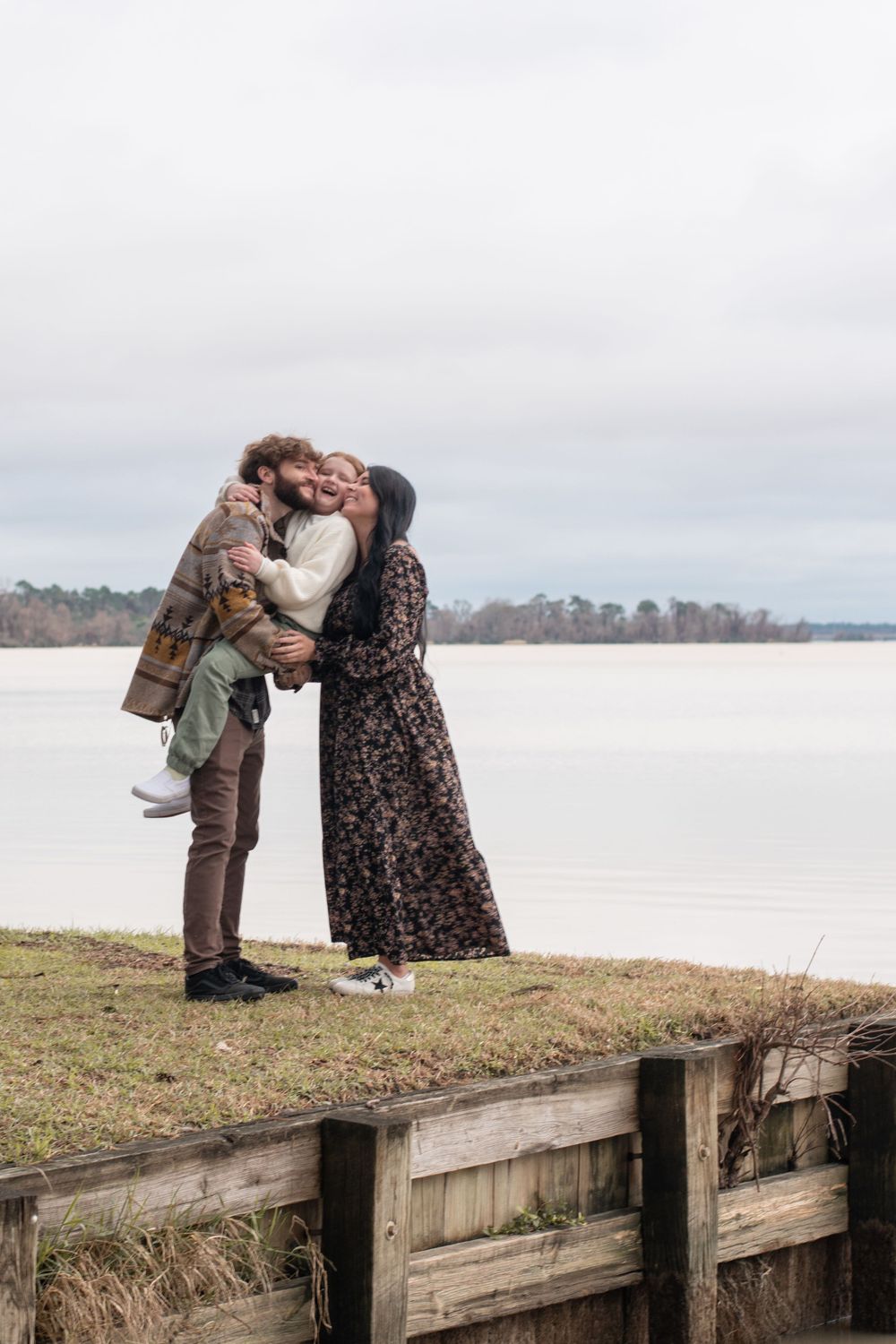 A couple shares a tender moment on a wooden pier overlooking a serene lake on an overcast day.