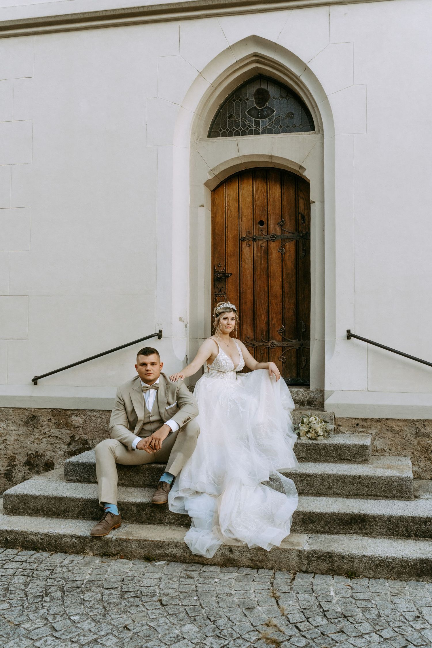 A couple in wedding attire sits playfully on stone church steps during a photo session.
