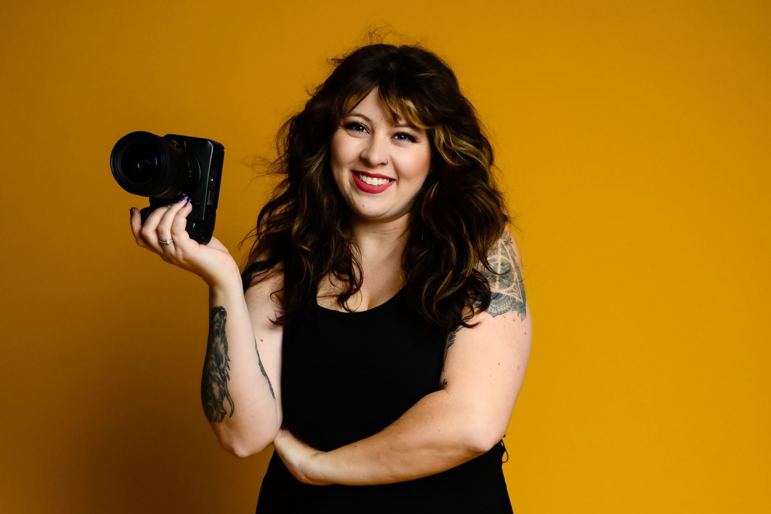 Female photographer smiling and holding a camera against a vibrant yellow background with tattoos visible.