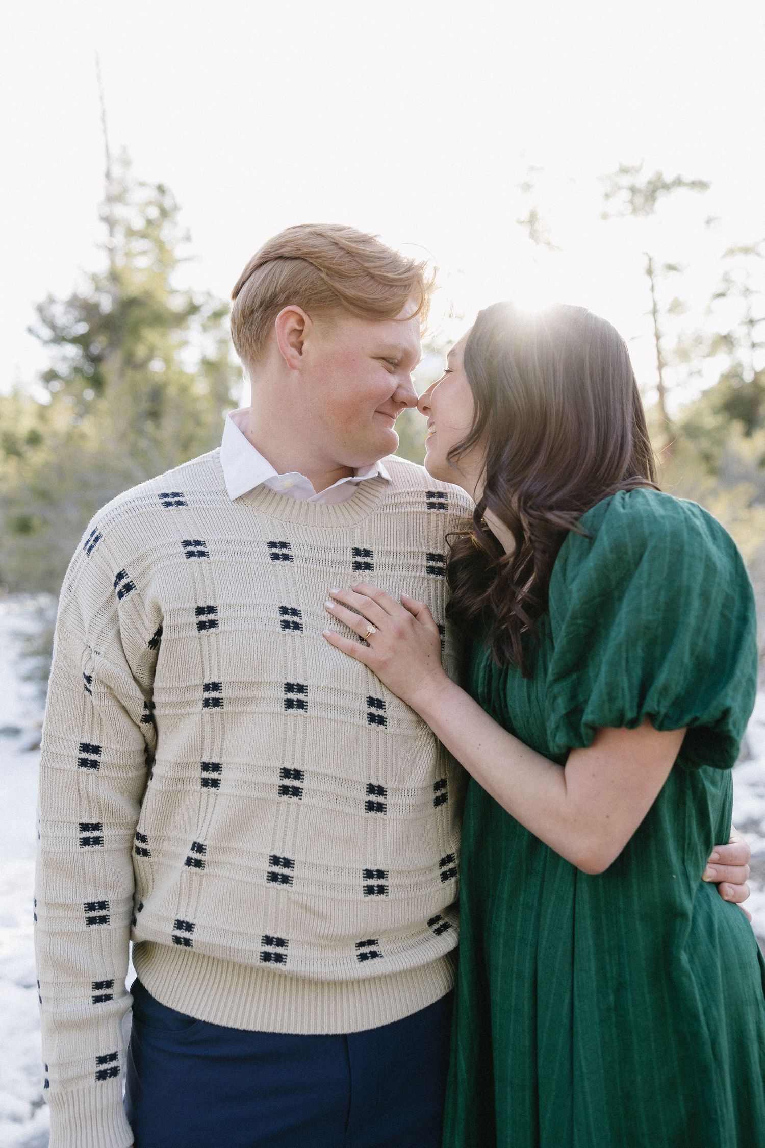 A romantic outdoor portrait of a couple in winter wear, with the person in green leaning lovingly against their partner.