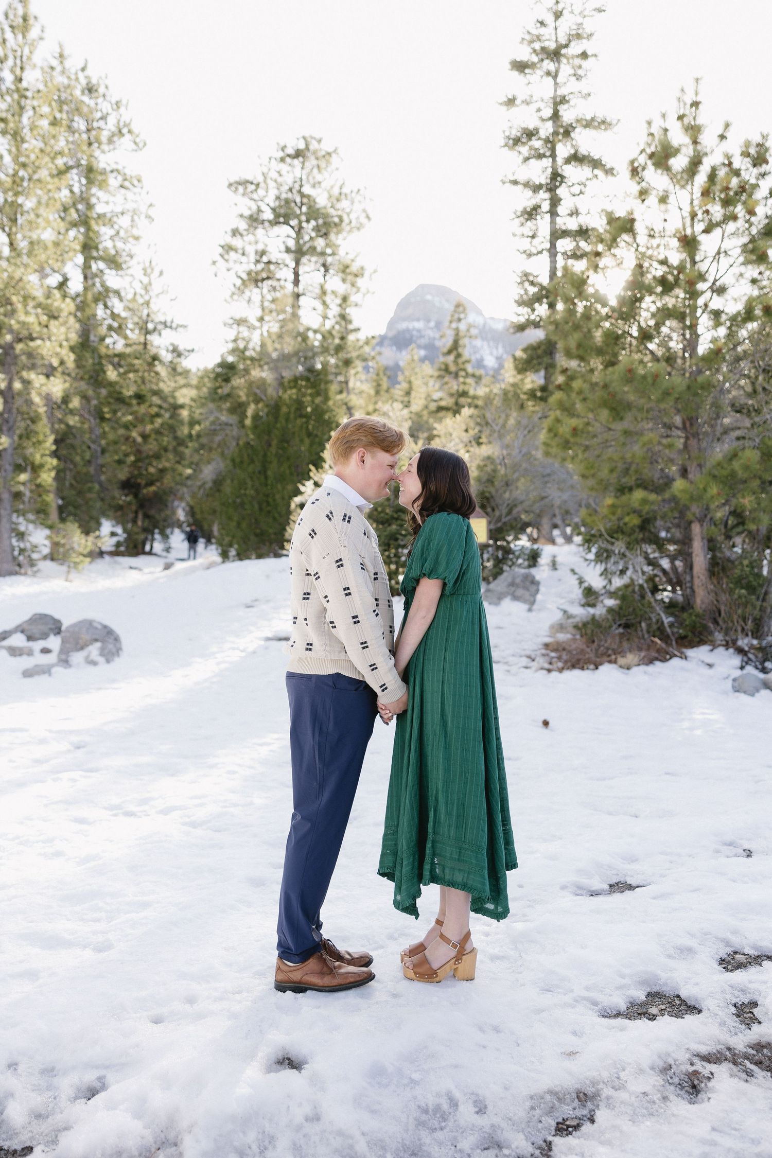 A couple embraces on a snowy mountain trail with evergreen trees and mountains in the background.