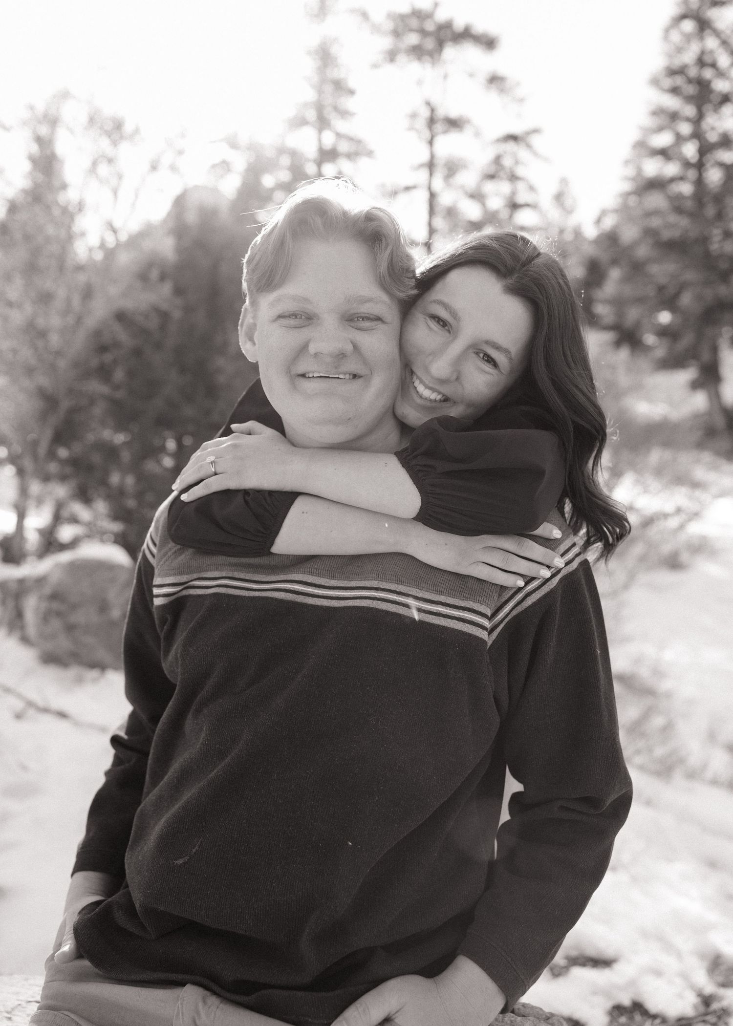A black and white portrait shows a couple embracing outdoors in winter, with pine trees visible in the background.