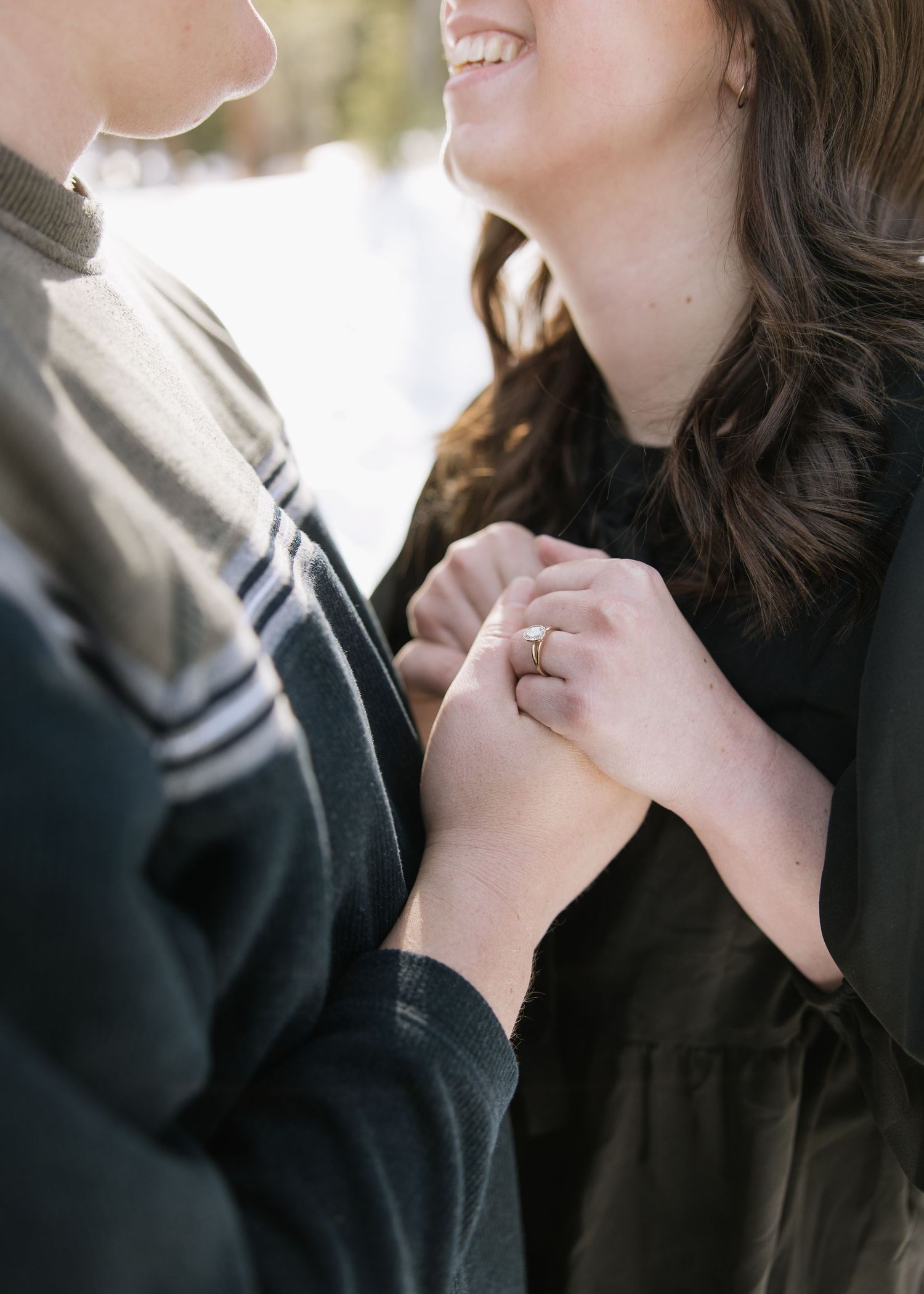 A joyful couple hold hands and smile during an intimate engagement moment with a ring visible.