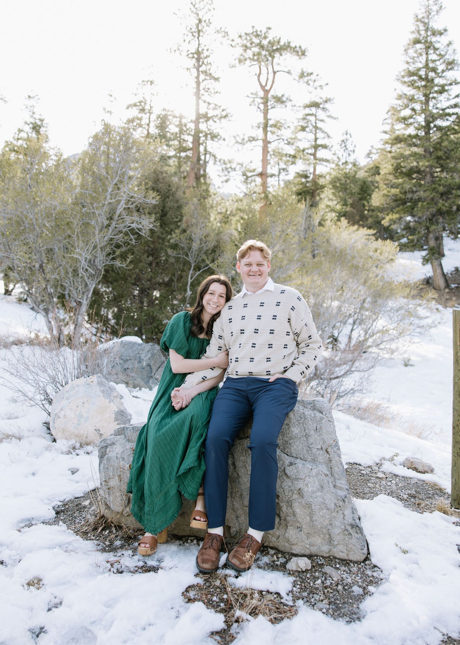 A couple sits together on a snow-covered boulder in a wintry mountain setting surrounded by evergreen trees.