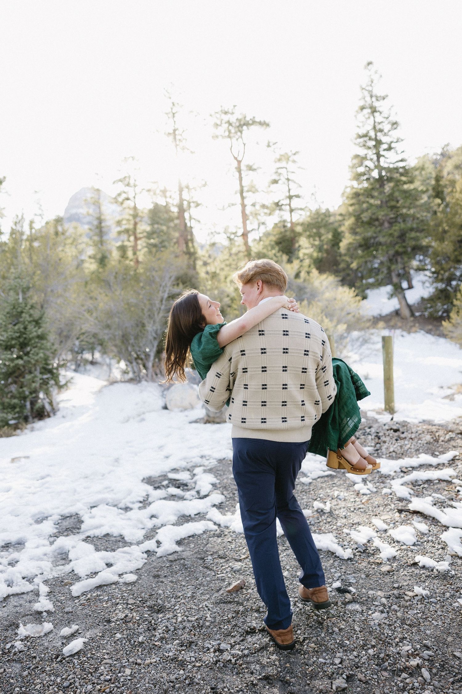 A couple shares a romantic moment in a snowy mountain landscape surrounded by evergreen trees on a winter day.