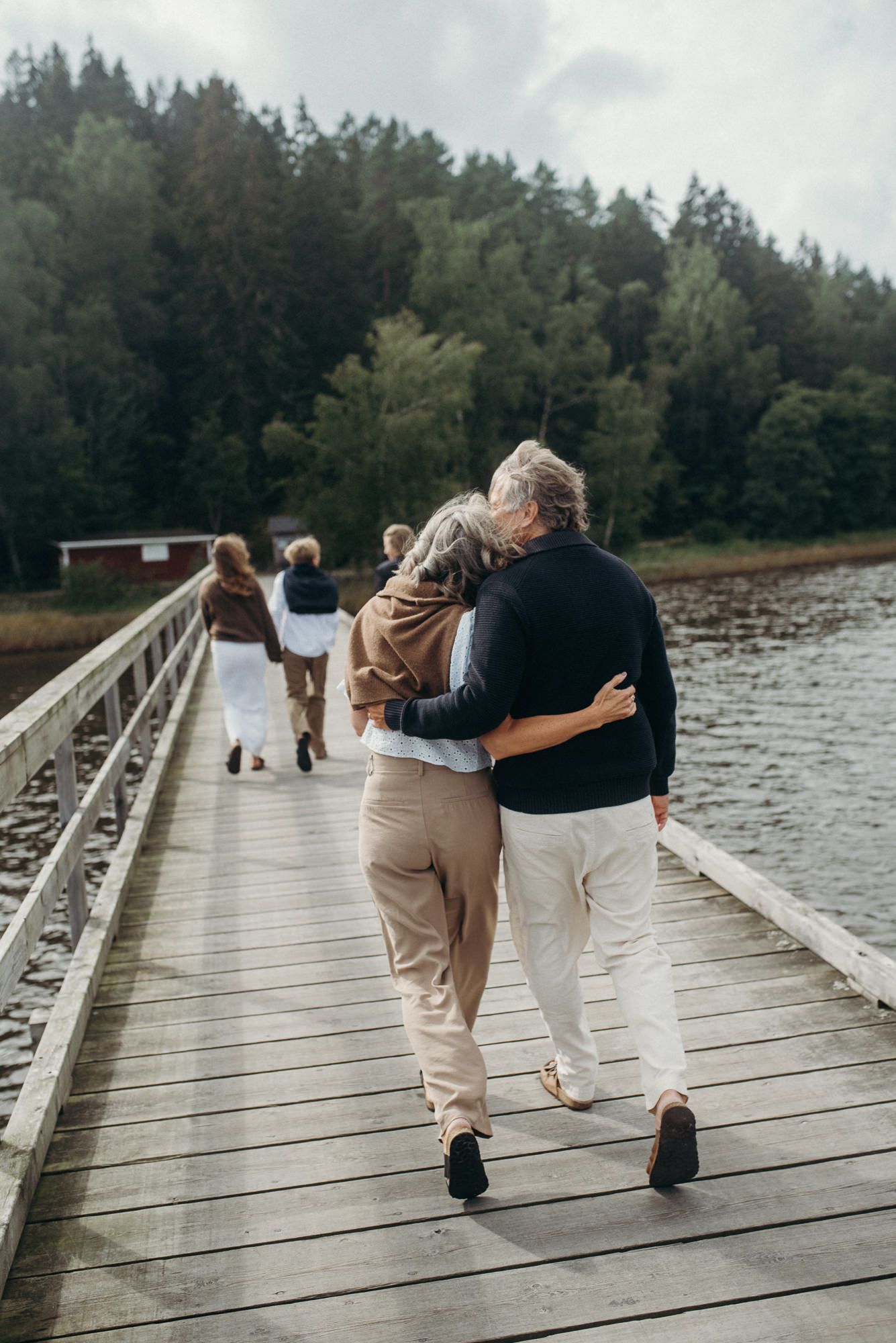 Couple walking arm in arm on wooden boardwalk by lake surrounded by evergreen forest.