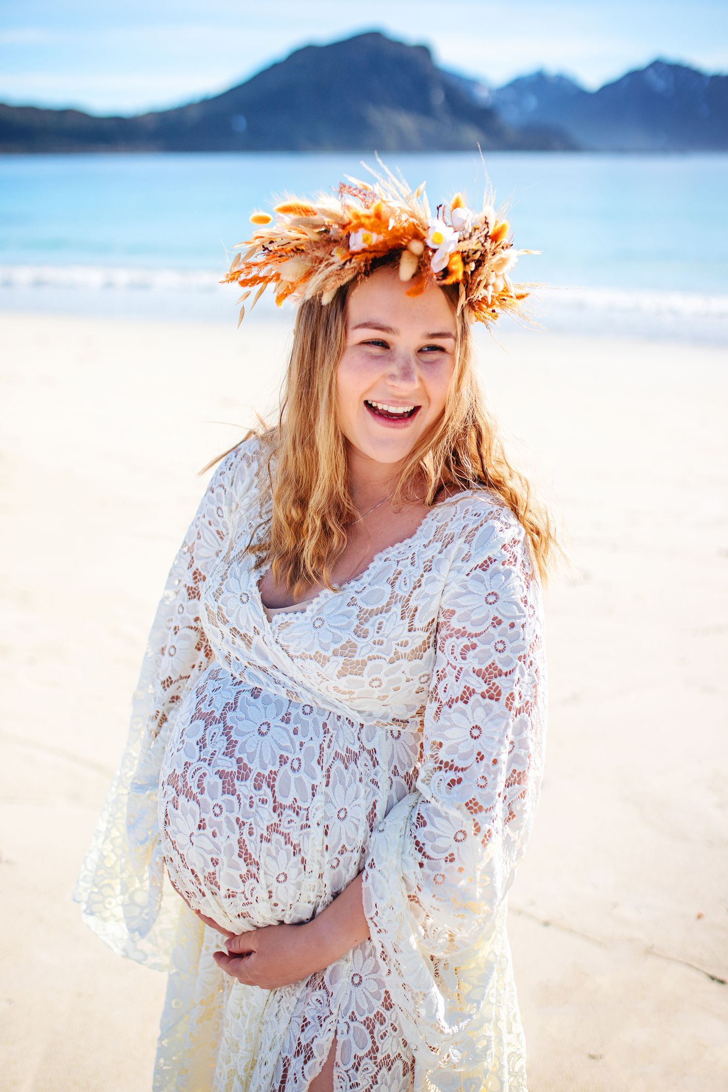 Laughing pregnant woman in white lace dress with autumn floral crown on sunny beach.
