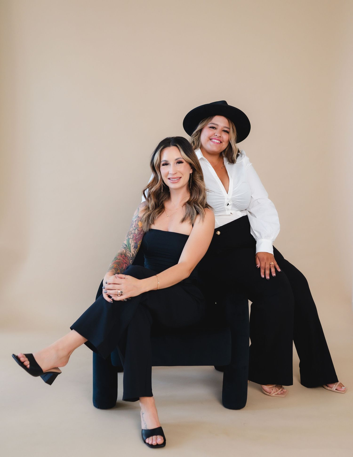 Two people in black and white outfits pose for a studio portrait sitting together on a chair against a beige backdrop.